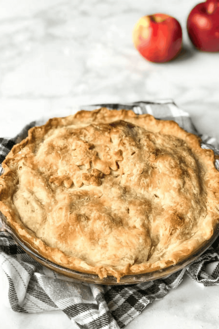 Easy homemade apple pie with a golden double crust and fluted edges, sitting on a plaid towel with fresh red apples behind it