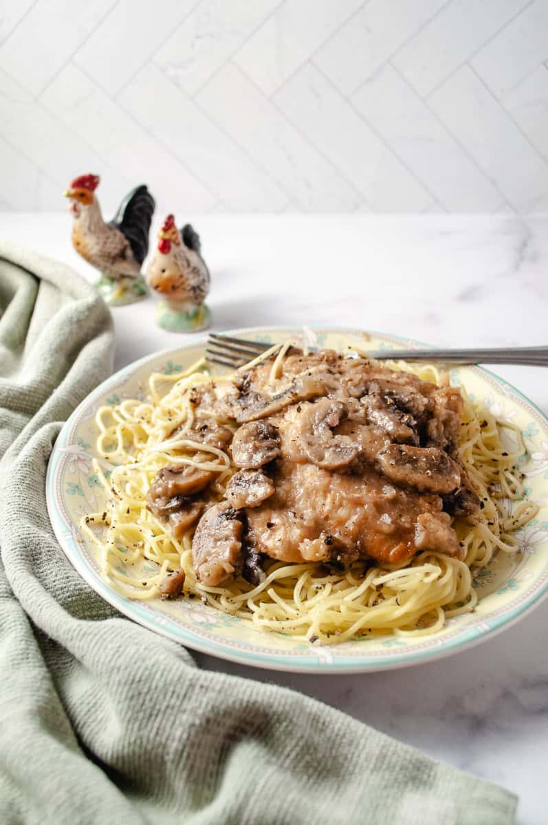 chicken marsala oven baked on a dinner plate on top of homemade pasta with a fork and 2 chicken salt and pepper shakers on a white counter a green towel on the counter wider view.