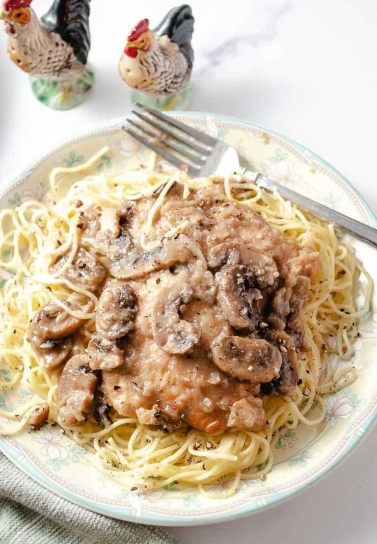 chicken marsala oven baked on a dinner plate on top of homemade pasta with a fork and 2 chicken salt and pepper shakers on a white counter.