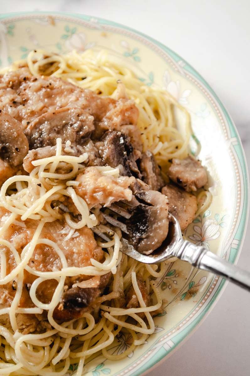 chicken marsala oven baked on a dinner plate on top of homemade pasta with a fork close up with the fork lifting up chicken, mushroom and pasta to eat.