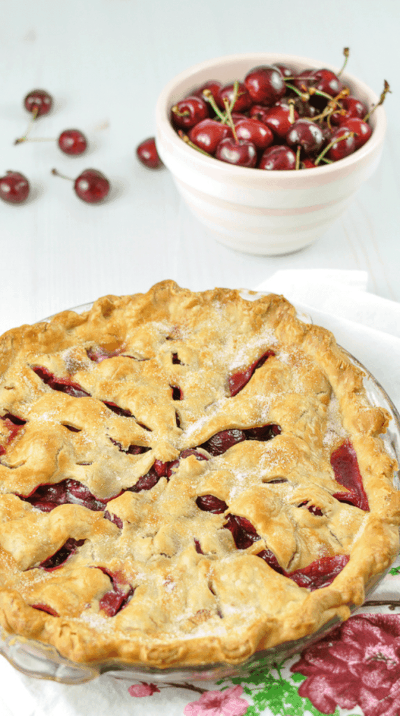 A baked cherry pie in a clear pie pan on a white counter with a white and pink striped bowl fold with cherries in the bathroom and cherries on the counter.