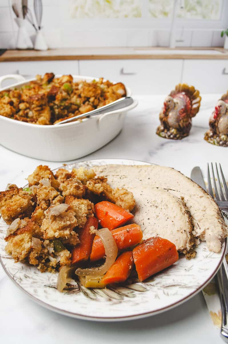  2 slices of turkey breast on a plate with cooked carrots and cornbread stuffing with a fork and knife on a white counter with a casserole of cornbread stuffing in the background and a turkey salt and turkey pepper shaker. 