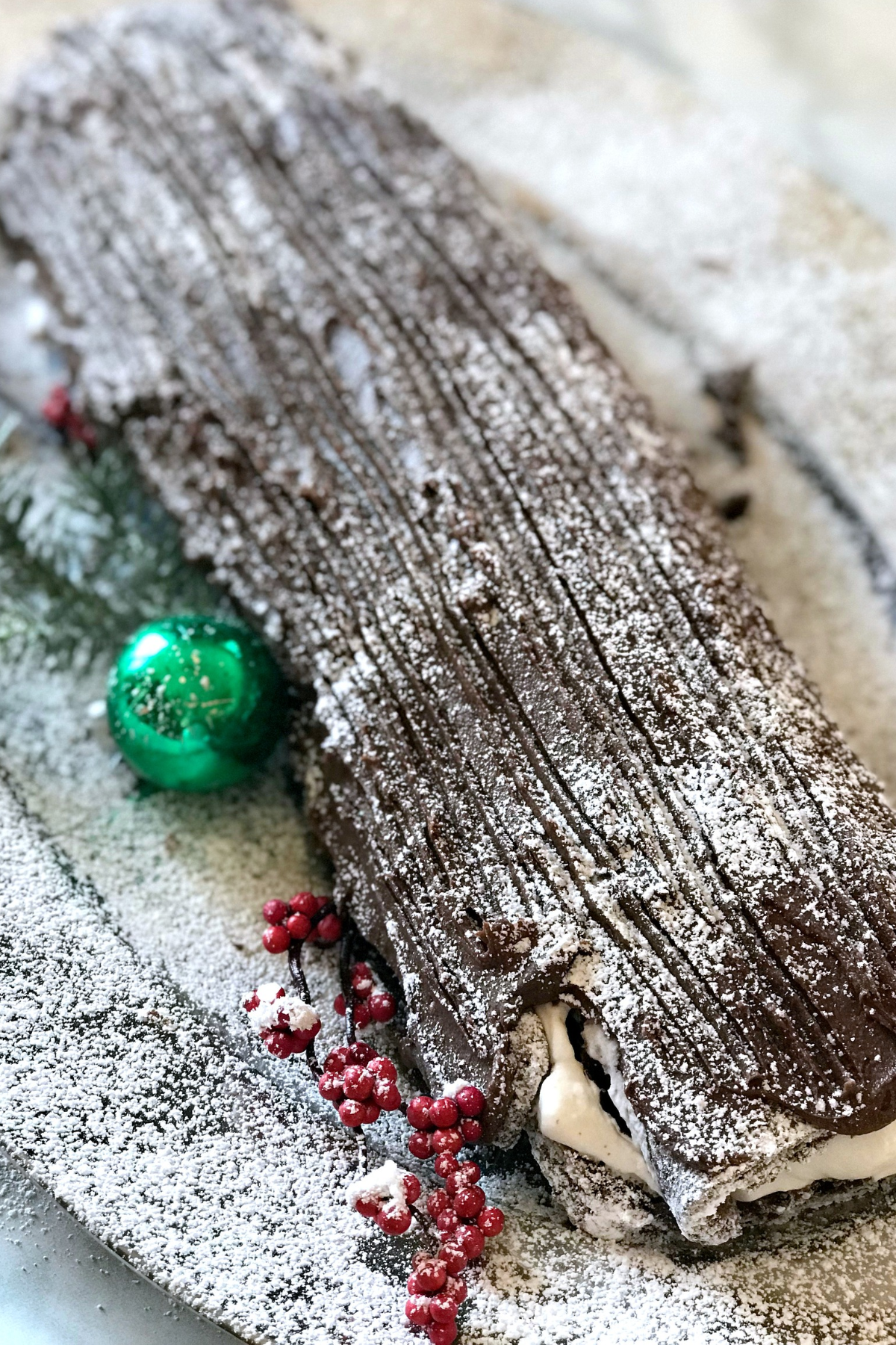 Christmas yule log cake recipe with chocolate bark frosting and powdered sugar snow, whipped cream filling showing at the end, a green Christmas bulb, and red berries on a platter