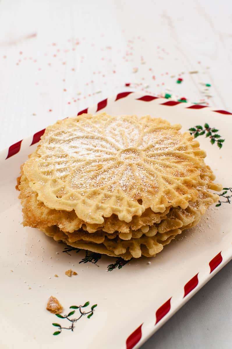 a stack of golden brown pizzelle cookies on a christmas plate with confectioners sugar on top. 
