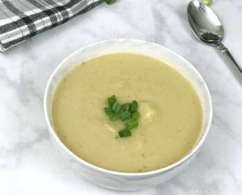 potato soup in a white bowl on a marble table with a spoon and a black checkered towel.