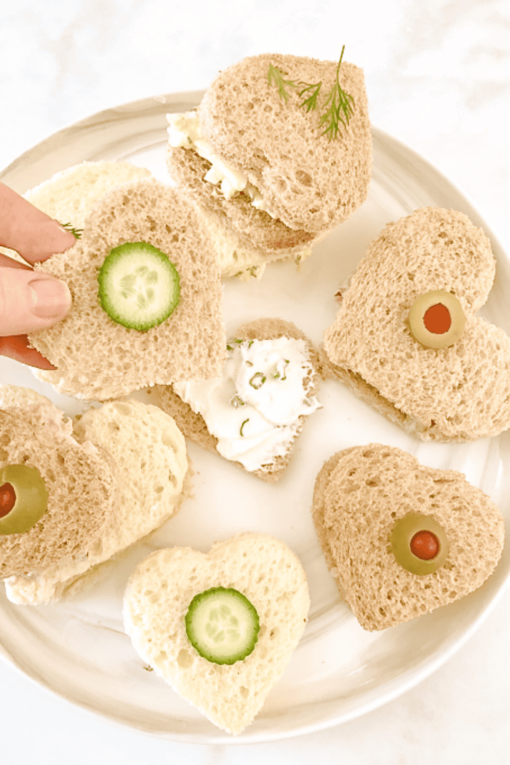 7 heart shaped tea sandwiches on a white plate some with mini cucumbers on top and some with slices of olives.