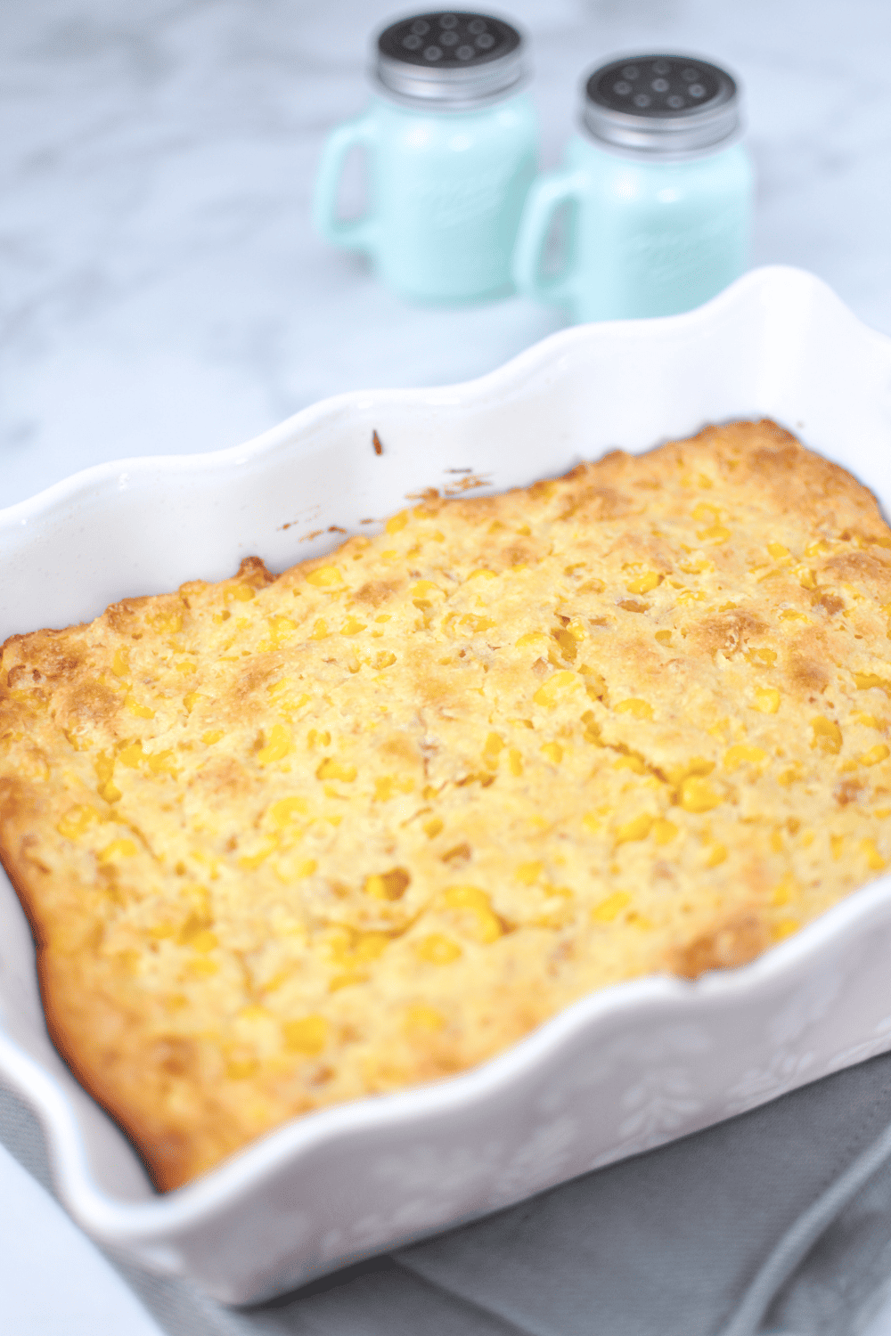 Corn Bake Jiffy cornbread mix in a pink casserole dish on a white counter with blue salt and pepper shakers in the background.