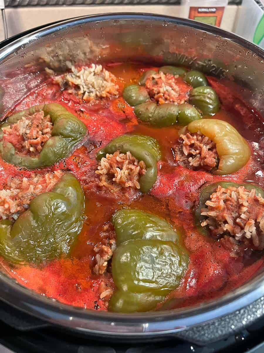 Several stuffed green peppers filled with ground beef and rice cook together in tomato sauce inside an Instant Pot insert sitting on the stovetop.