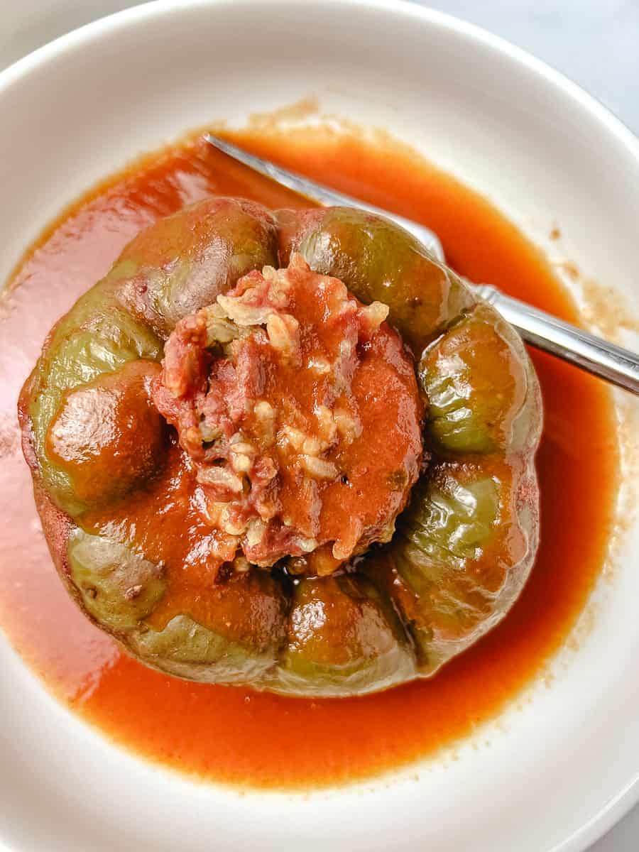 An overhead view of a stuffed green pepper with ground beef and rice sitting in a white bowl of tomato sauce, with a fork resting against the side of the bowl.