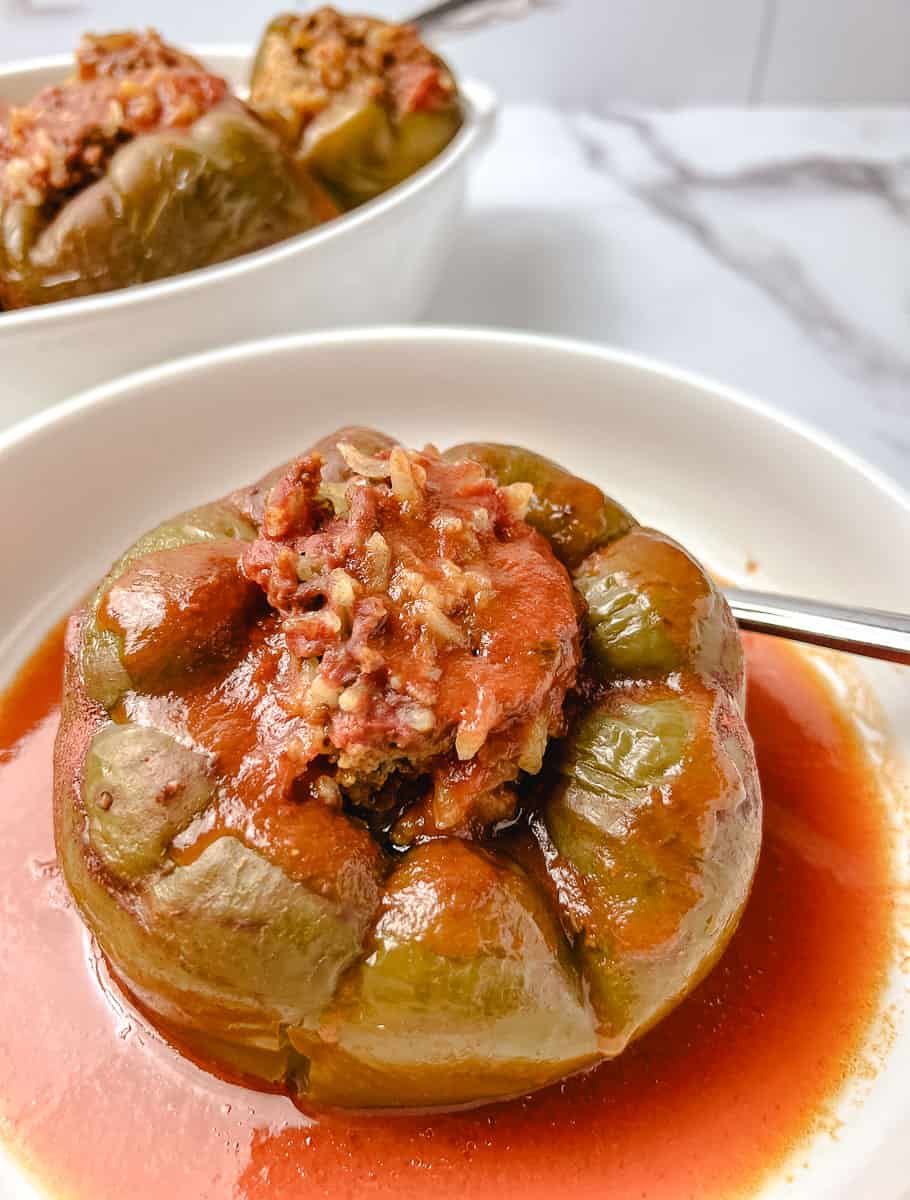 Close up of a stuffed green pepper recipe made with ground beef and rice, topped with tomato sauce, served in a white bowl with a spoon resting beside it.