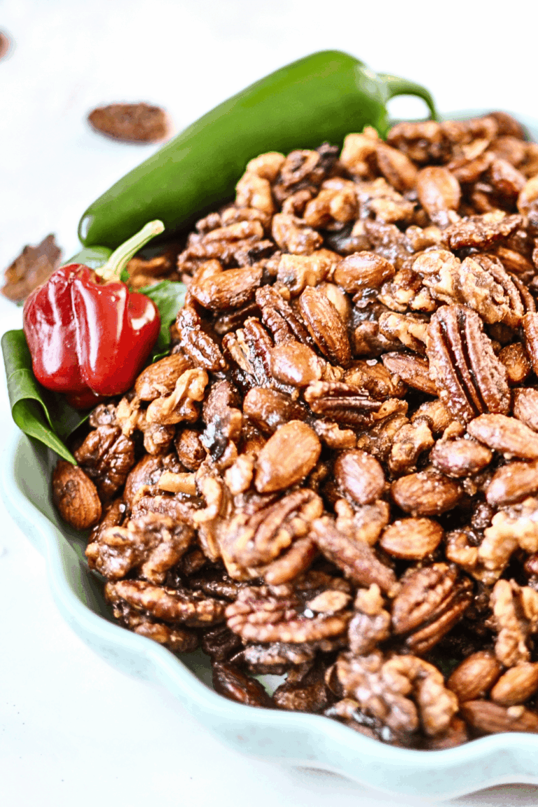 Christmas spiced nuts, a glossy mix of pecans, almonds, and walnuts, piled in a mint green scalloped dish with a fresh jalapeño and a small red pepper on a white marble counter.