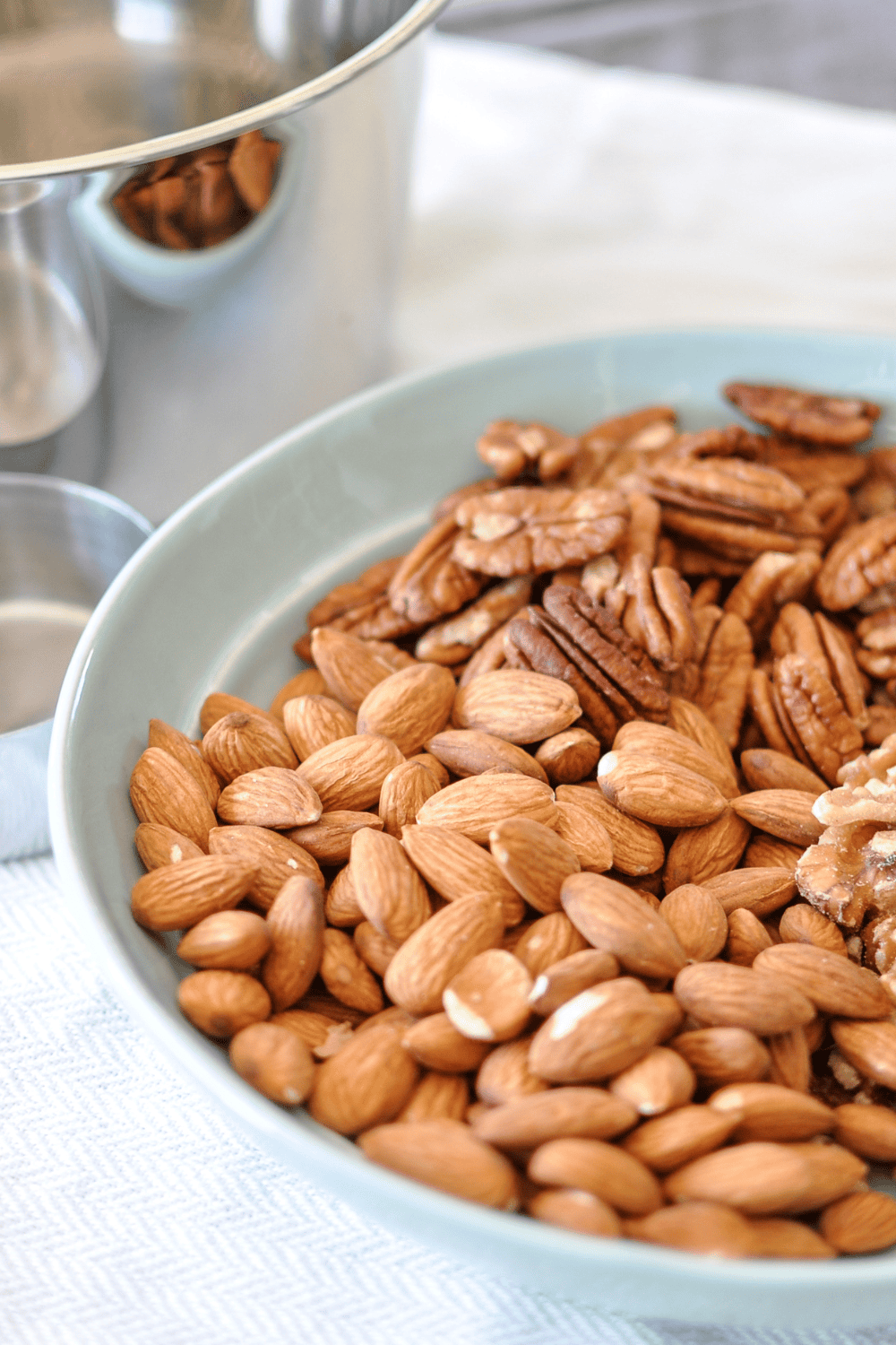 Raw mixed nuts for Christmas spiced nuts, almonds, pecans, and walnuts, in a pale blue bowl on a herringbone linen with a metal mixing bowl behind.