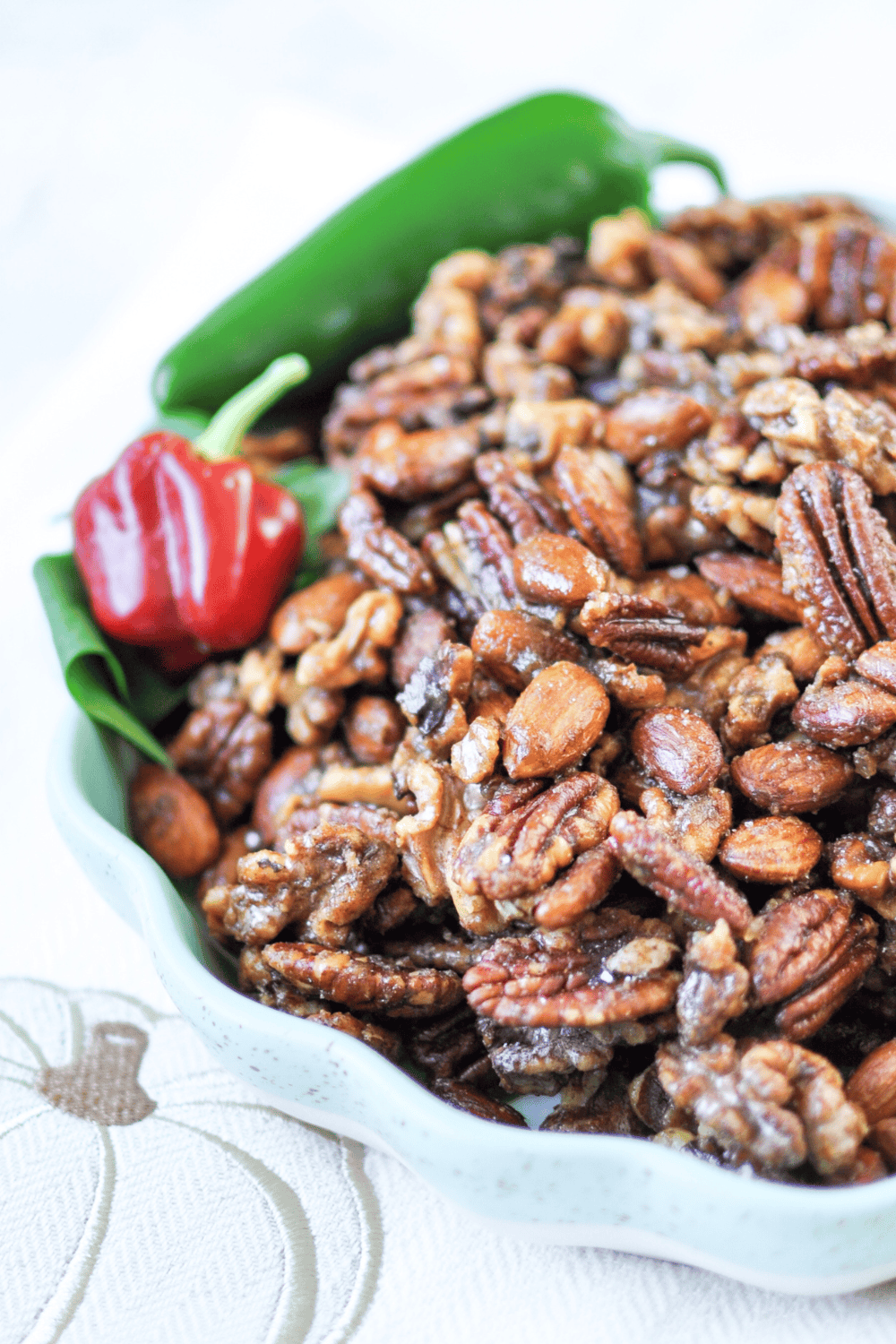 Christmas spiced nuts, glossy pecans, almonds, and walnuts, in a white scalloped dish with a green jalapeño and a red pepper, resting on a pumpkin print napkin.