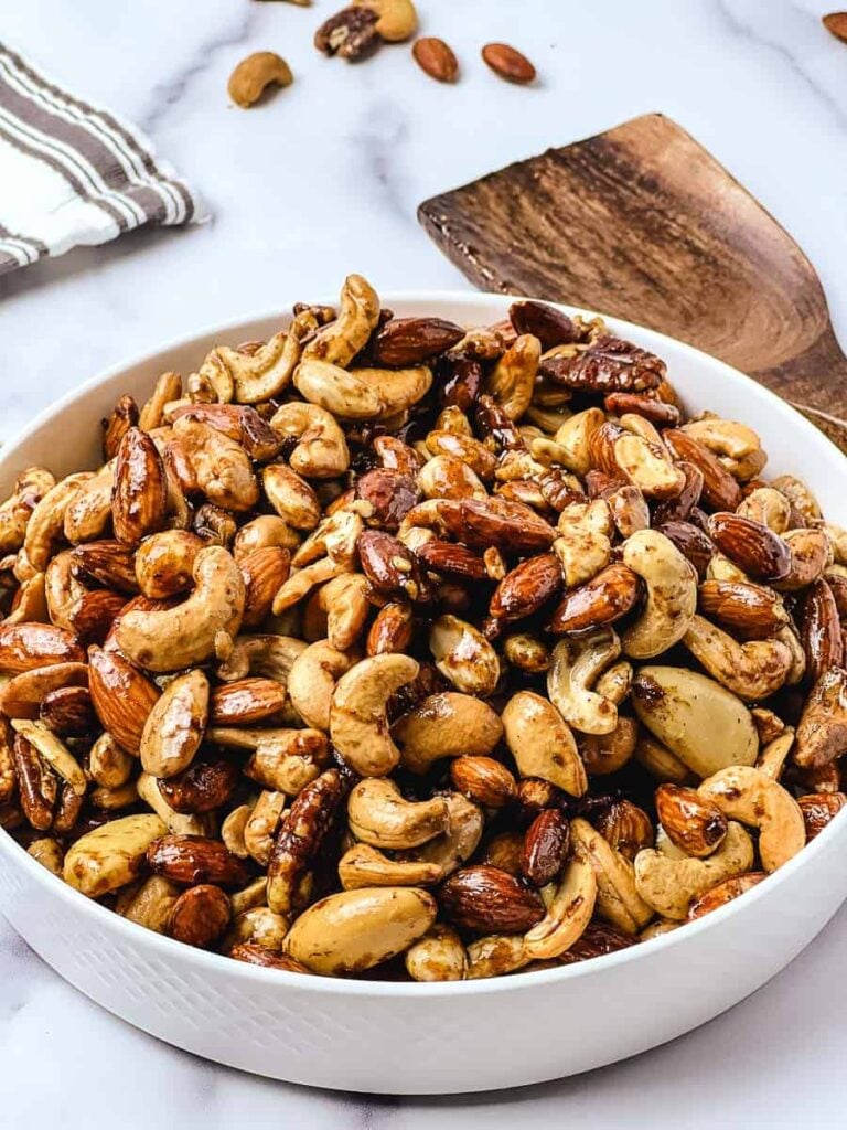 Christmas spiced nuts piled in a white bowl on a marble counter with a wooden spatula and striped towel