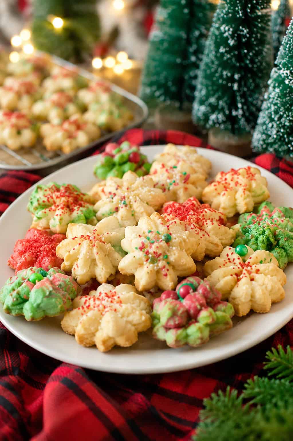 Christmas spritz cookies on a white plate with red and green sprinkles, surrounded by holiday decorations and fresh baked cookies in the background.