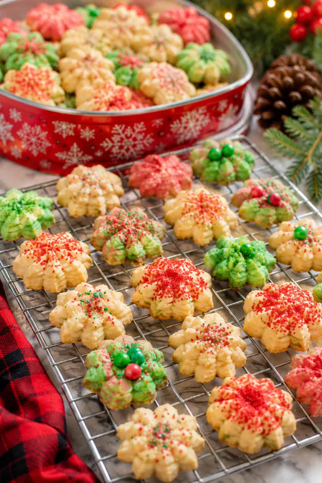 Christmas spritz cookies on a white plate with red and green sprinkles, surrounded by holiday decorations and fresh baked cookies in the background.