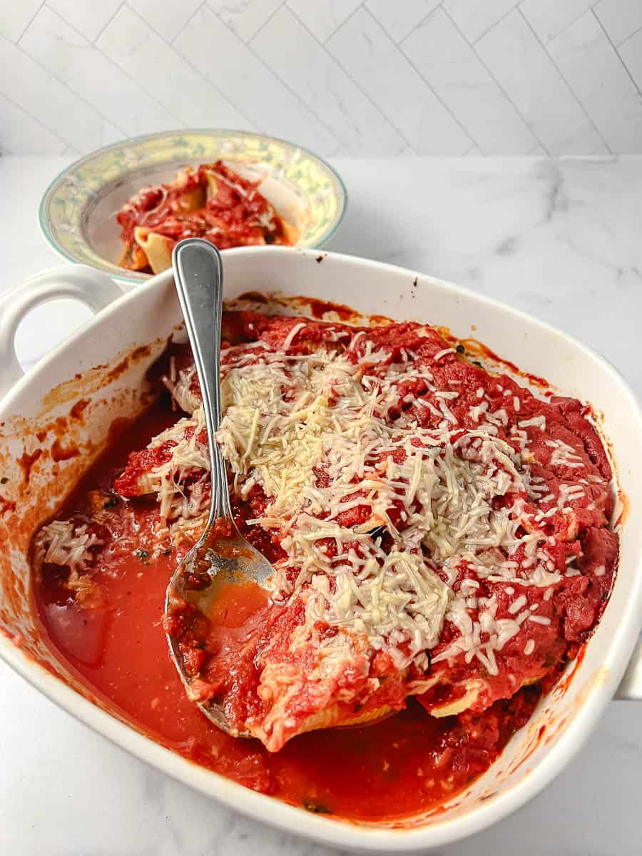 Easy Vegetarian Stuffed Shells Spinach and Ricotta Cheese 3 in a white square baking dish on a marble counter with a serving of three shells in the background and a white bowl with yellow and green flower and trim.