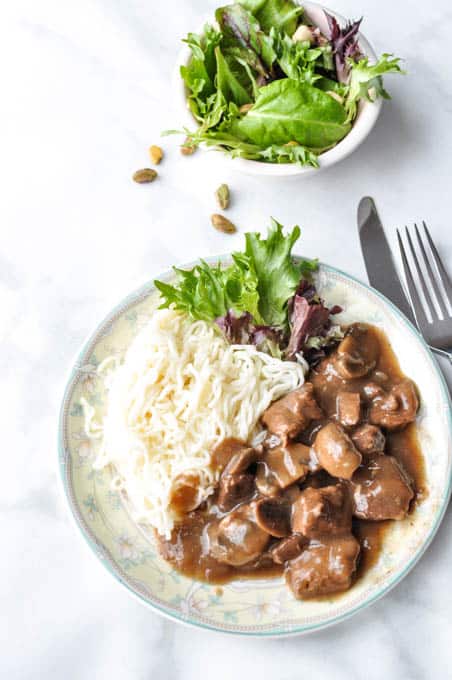 Instant Pot Beef Tips With Mushroom Gravy on a white plate with mashed potatoes and salad,  small plate of salad in the background and a fork and a knife. 