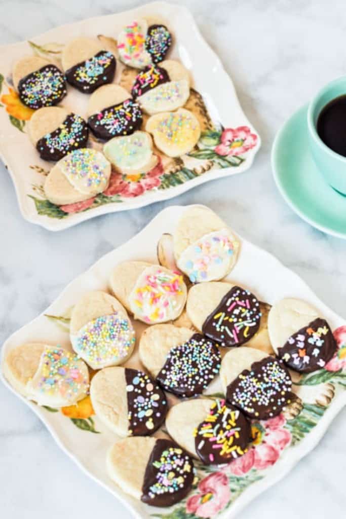 Quick sugar cookies dipped in chocolate with sprinkles arranged on a white plate with a cup of coffee