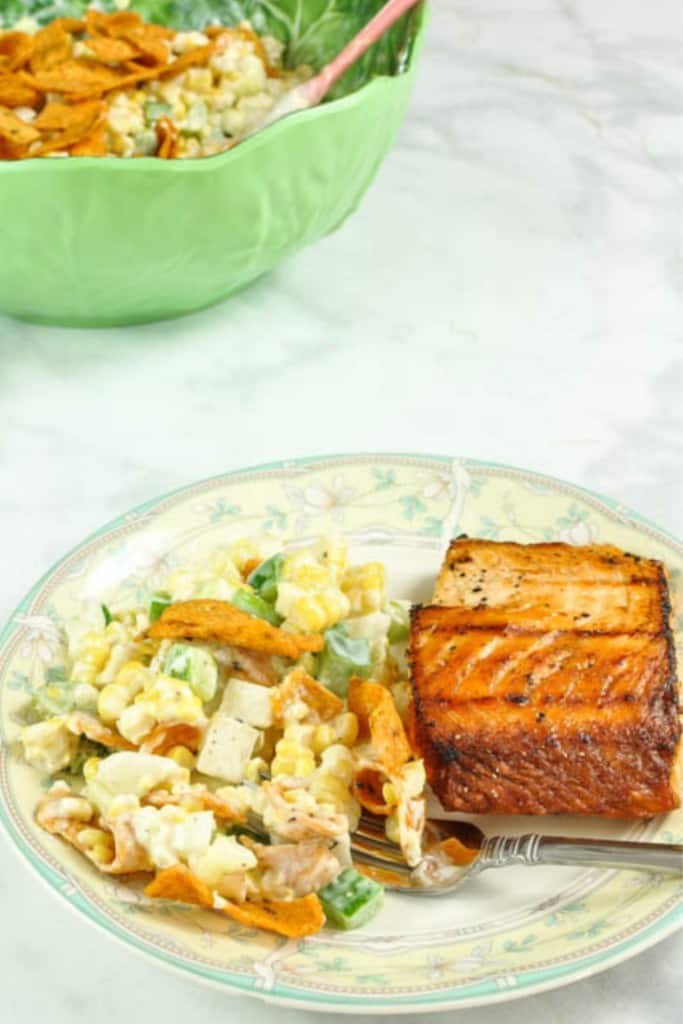 Easy Spicy Frito Corn Salad with Cheese on a yellow and green flowered rimmed plate with a slice of salmon and a fork with a green serving bowl in the background with the corn salad in it.