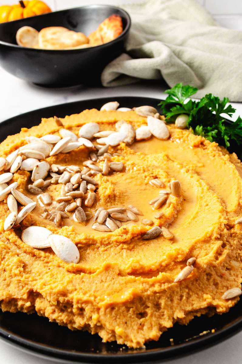 close up pumpkin hummus on a black plate with pumpkin seed on top parsley on side of the plate top of image a black bowl with little bread dippers a mini pumpkin and a green towel.