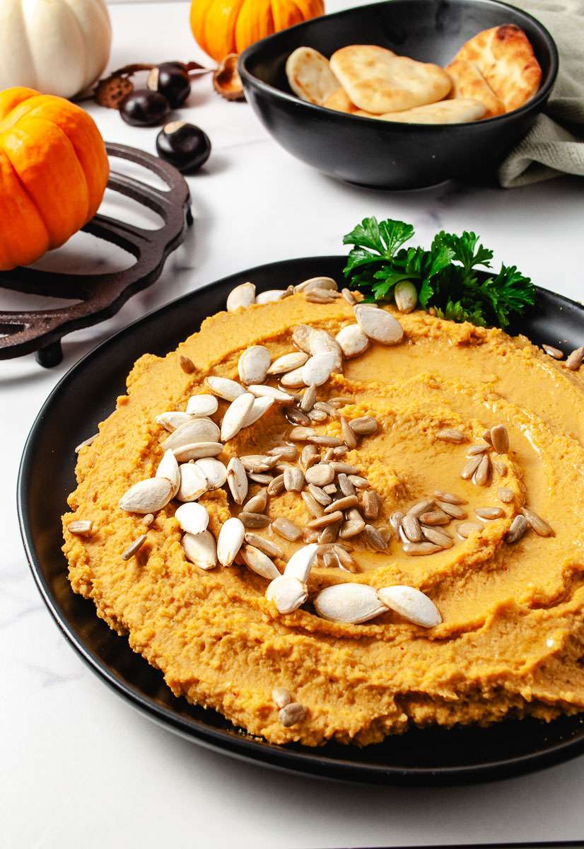 pumpkin hummus on a black plate with pumpkin seed on top parsley on side of the plate top of image a black bowl with little bread dippers a 3 mini pumpkin and a green towel with some buckeye nuts and one pumpkin on a pumpkin trivit.