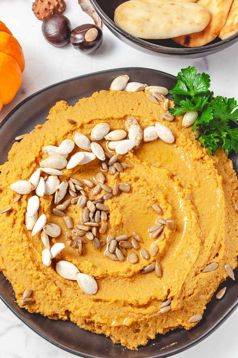 close up top downpumpkin hummus on a black plate with pumpkin seed on top parsley on side of the plate top of image a black bowl with little bread dippers a mini pumpkin 2 buckeye nuts on a white counter