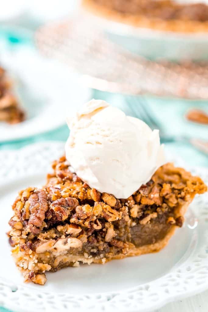 a lice of pecan pie on.a white plate with a scoop of vanilla ice cream on top and the whole pie in the back ground. 