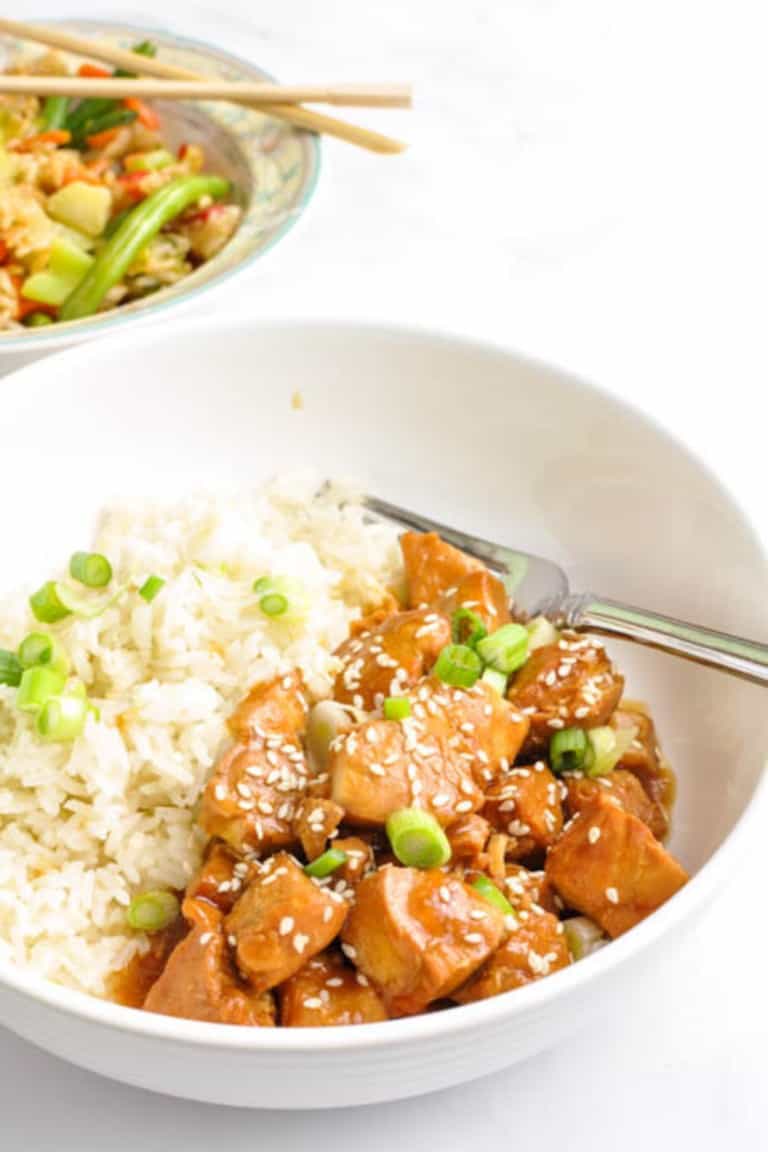 Instant Pot Honey Sesame Chicken Breast Anna White bowl Anna White counter with rice and green onions on top a fork in the bowl and at the top left a bowl of vegetables with chopsticks.