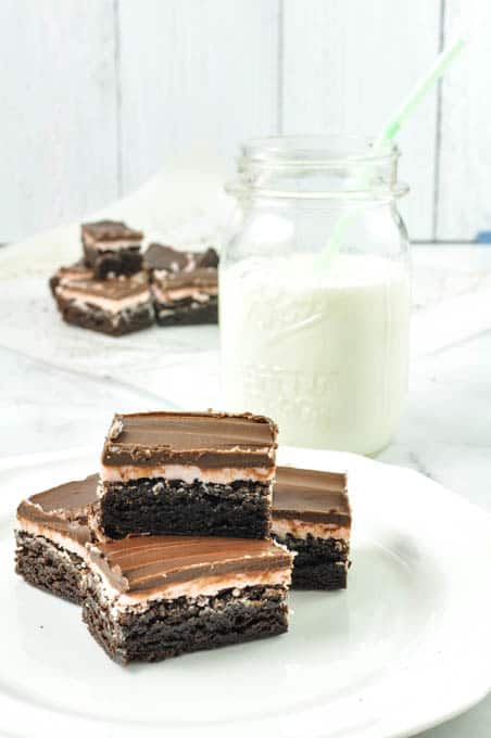 Best Mint Frosted Brownie recipe made easy and baked to fudgy a double layer of min frosting and chocolate ganache on a white plate with a mason jar of milk and a straw and more brownies in the background.