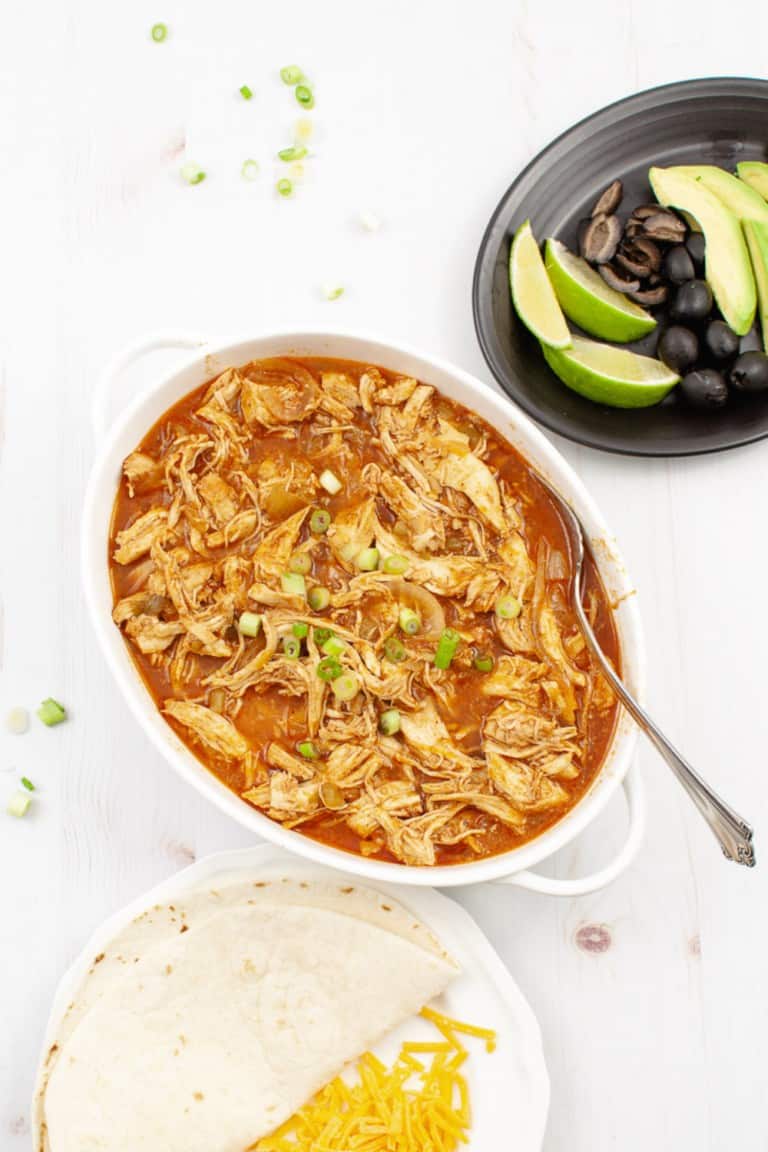 Crock Pot Enchilada Chicken Anna White serving bowl top view with a quesadilla and a white plate in front and a black plate with limes avocado and black olives.