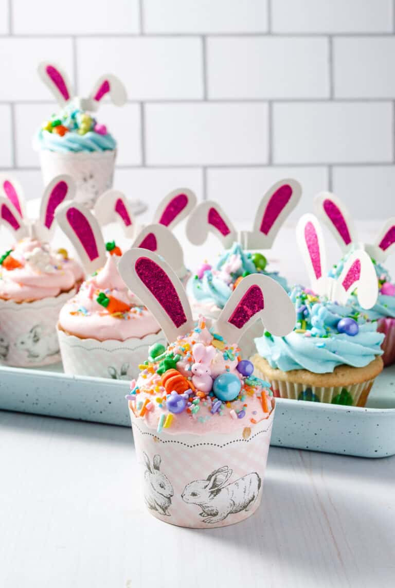 Bunny Ear Easter Cupcakes 8 some with pink frosting and some with blue, multi color sprinkes with white bunny ears with pink sparkle ears sticking form the top, on a blue baking tray on a white counter with a white subway tile background