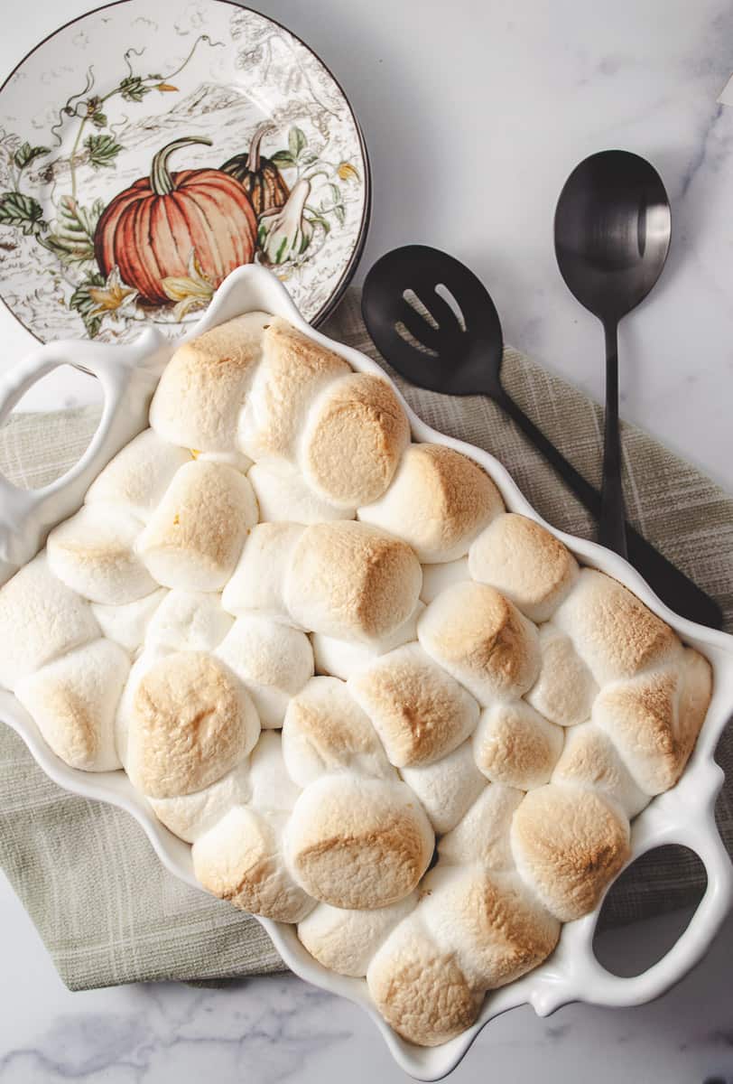 Candy sweet potato casserole cooked with golden brown toasted marshmallows on top with two black mat serving spoons and a small plate with a pumpkin design top down view.