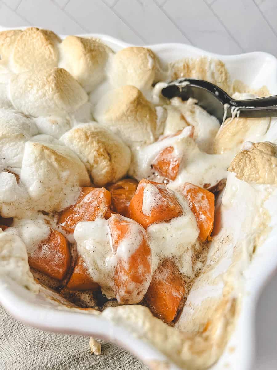 Candied sweet potato casserole close-up of the sweet potatoes with green melted marshmallows on top and black serving spoon in the dish.