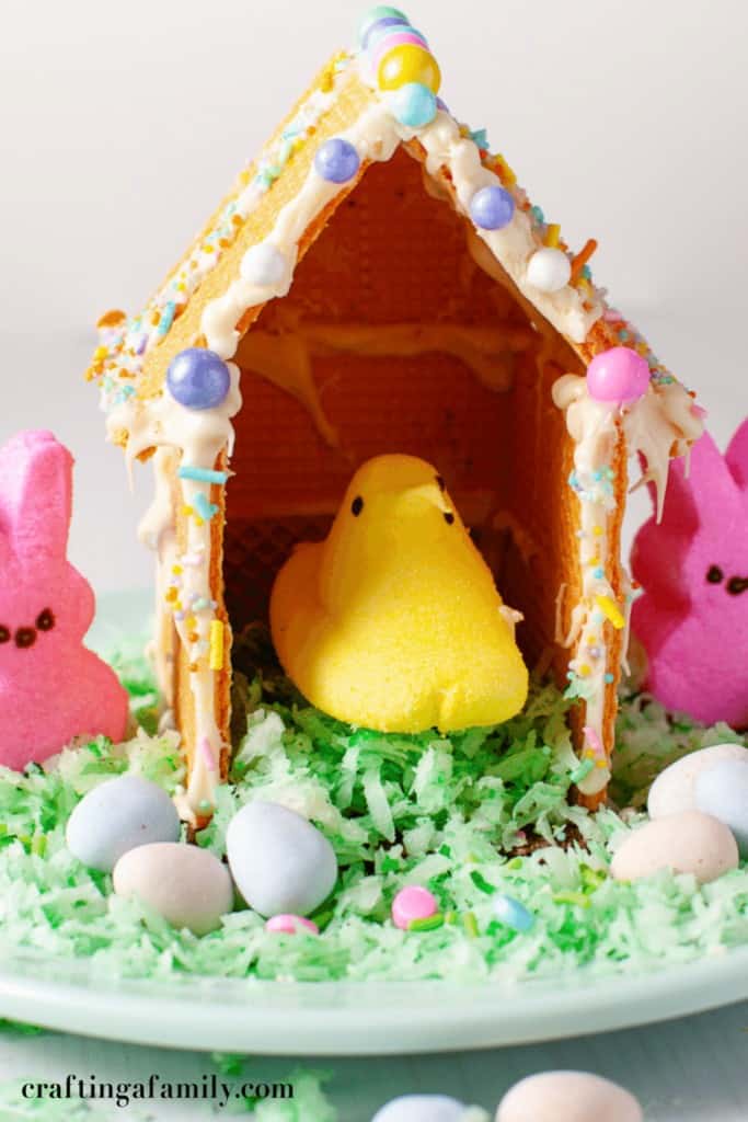 Cute Easter Peeps house kids craft made with graham crackers white icing pink bunny Peeps and a yellow chick surrounded by pastel candy eggs and green coconut grass