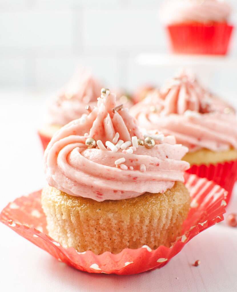 3 vanilla cupcakes with strawberry frosting on white table one cupcake with the red wrapper open