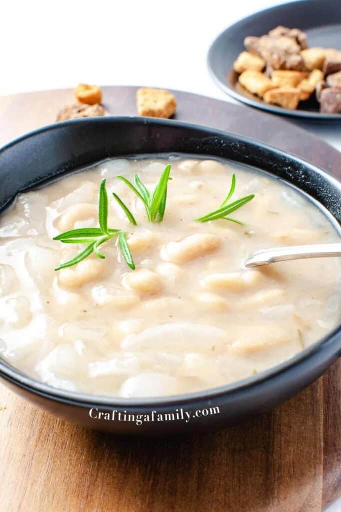 Vegan rosemary white bean soup in a black bowl with a spoon and a sprig of rosemary on a wooden platter with croutons