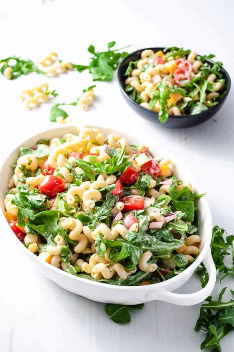 arugula pasta salad with cherry tomatoes in a white serving bowl and a small black bowl with a serving in the background.