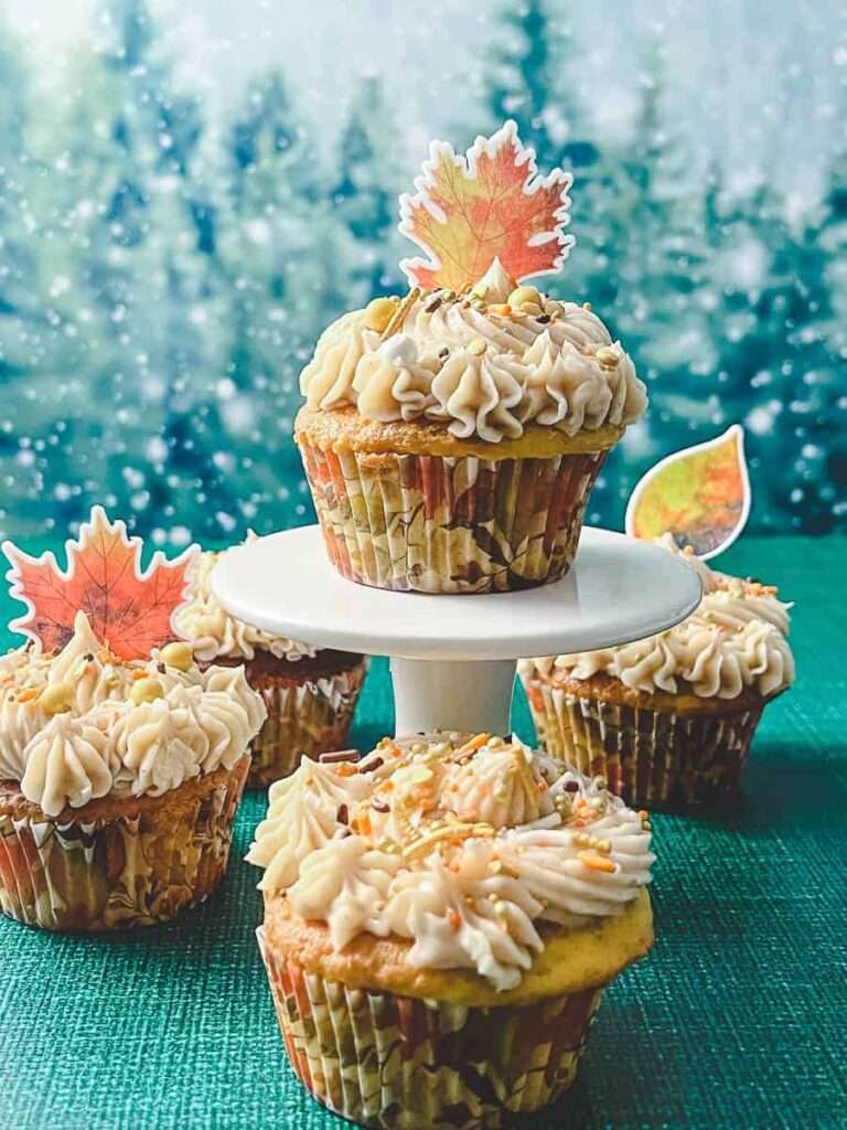 Apple cider fall cupcakes 4 on a blue green tablecloth one on a white pedestal with fall colored sprinkles and edible leaf candies on top.