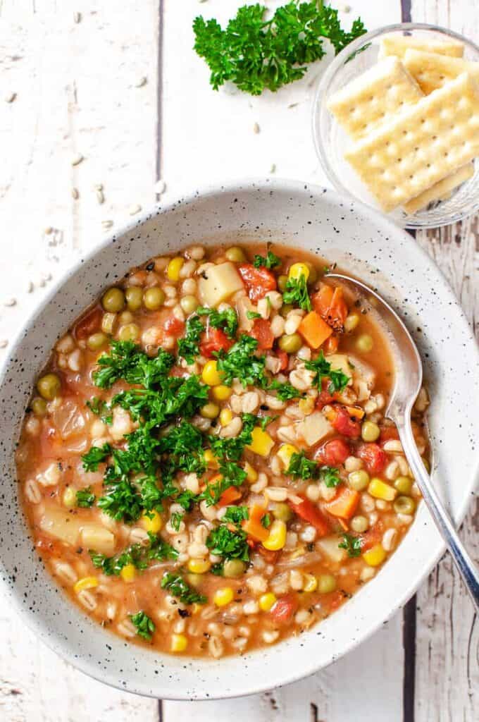 a gray bowl of barley soup with parsley on top and a spoon in the some parsley on the white wood table.