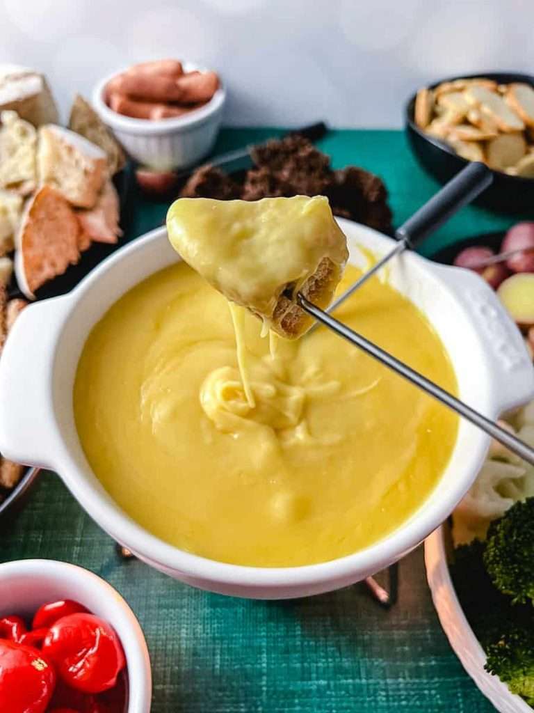 cheese sauce in white bowl on green table with bread and veggies, bread dipped in cheese on fork
