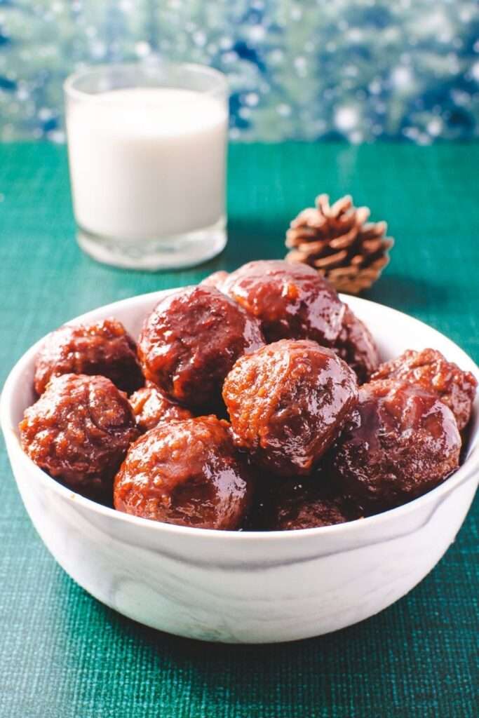 meatballs in grape jelly and chilli sauce in a white bowl on a green tablecloth and a white candle on the table. 