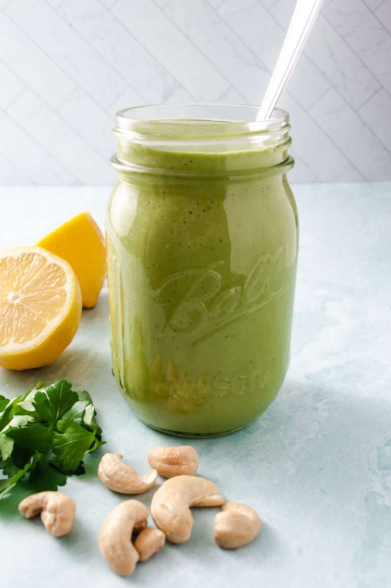 lemon cashew green goddess dressing no oil in a mason jar with spoon, cashews parsley and lemon on the counter. 