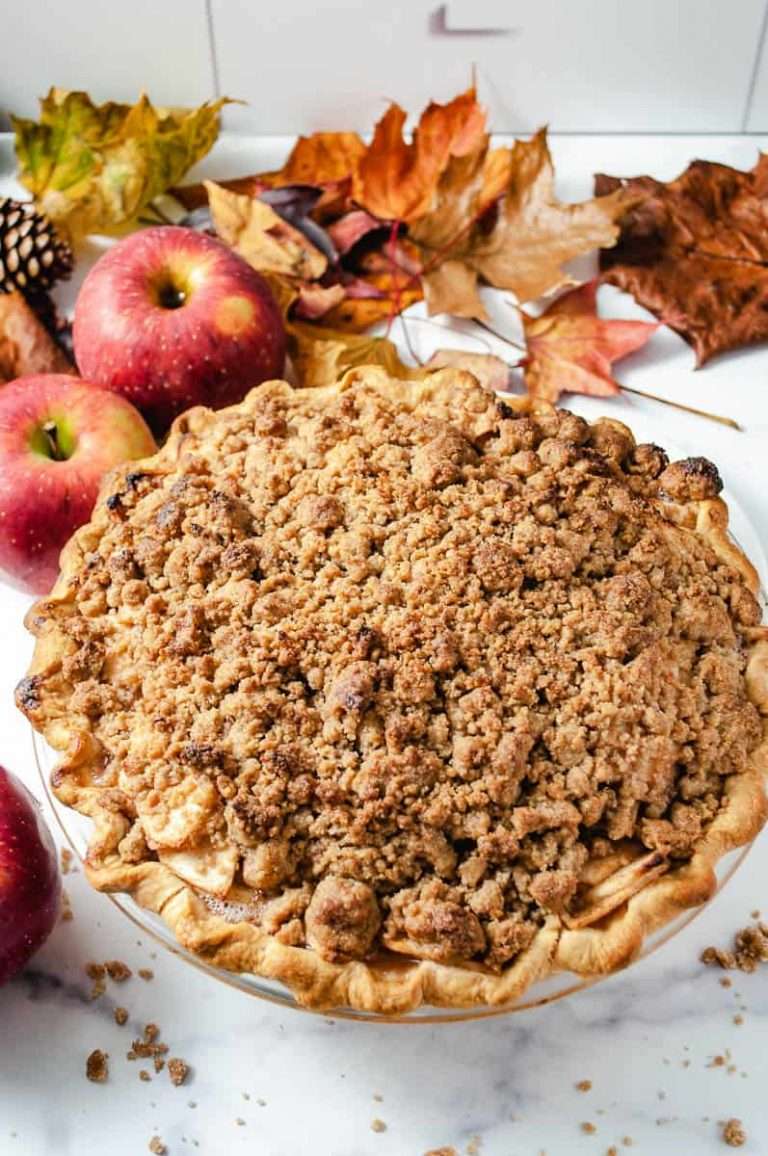 a whole apple crumble pie on a white counter and red apples and fall leaves on the counter.
