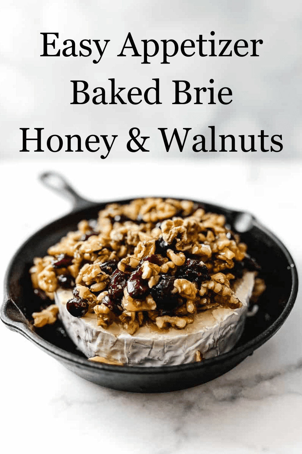 close up of honey being poured onto brie cheese in cast iron skillet with walnuts dried cranberries and walnuts on a white counter