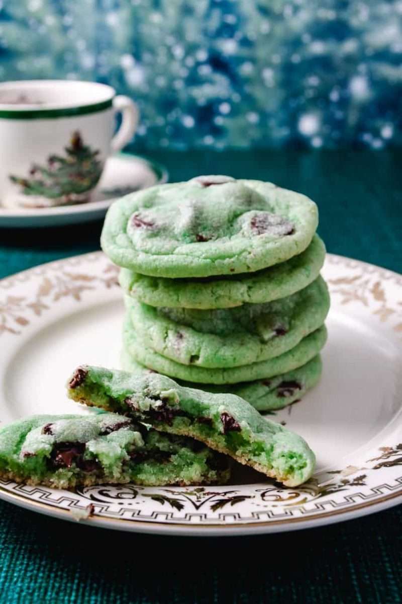 Stack of green mint chocolate chip cookies made with sugar cookie mix, displayed on a white and gold holiday plate. One cookie is broken in front to show the melty chocolate chips inside. A festive cup with a Christmas tree design and a snowy blue background set the holiday mood.