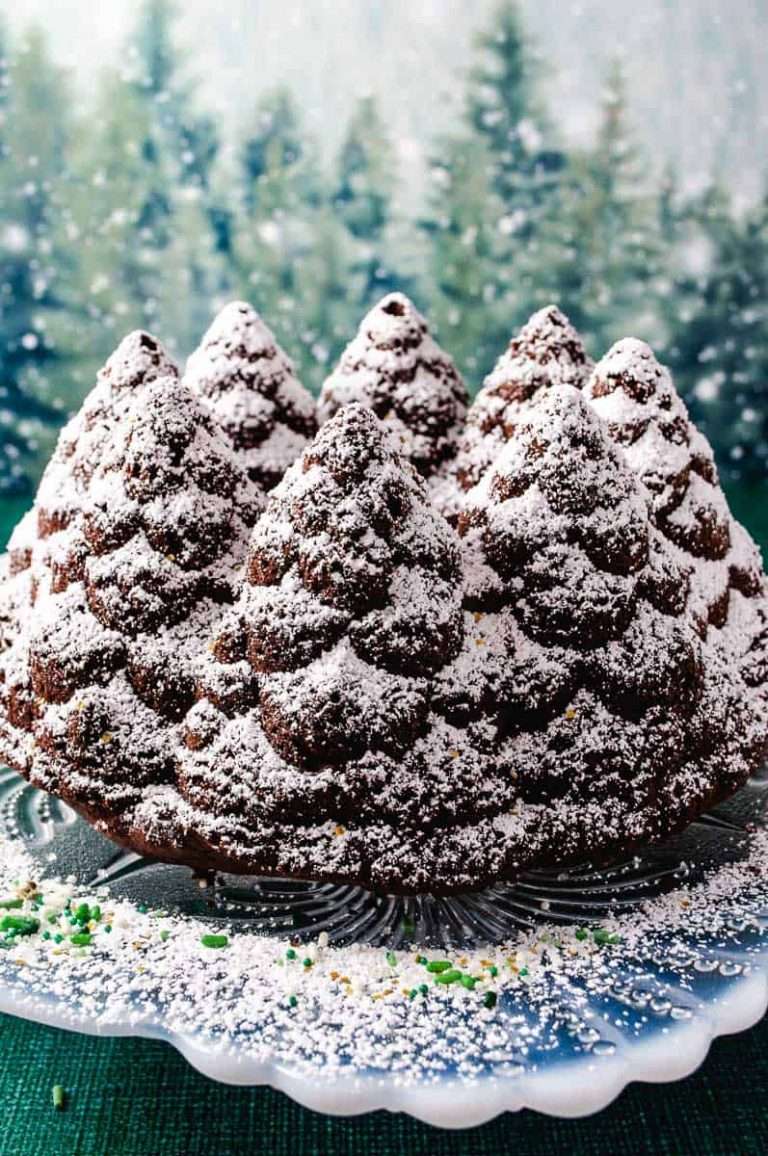 Chocolate Bundt Cake Recipe Pine Forest NordicWare Pan on a white glass cake stand with powdered sugar on a green table with pine forest background