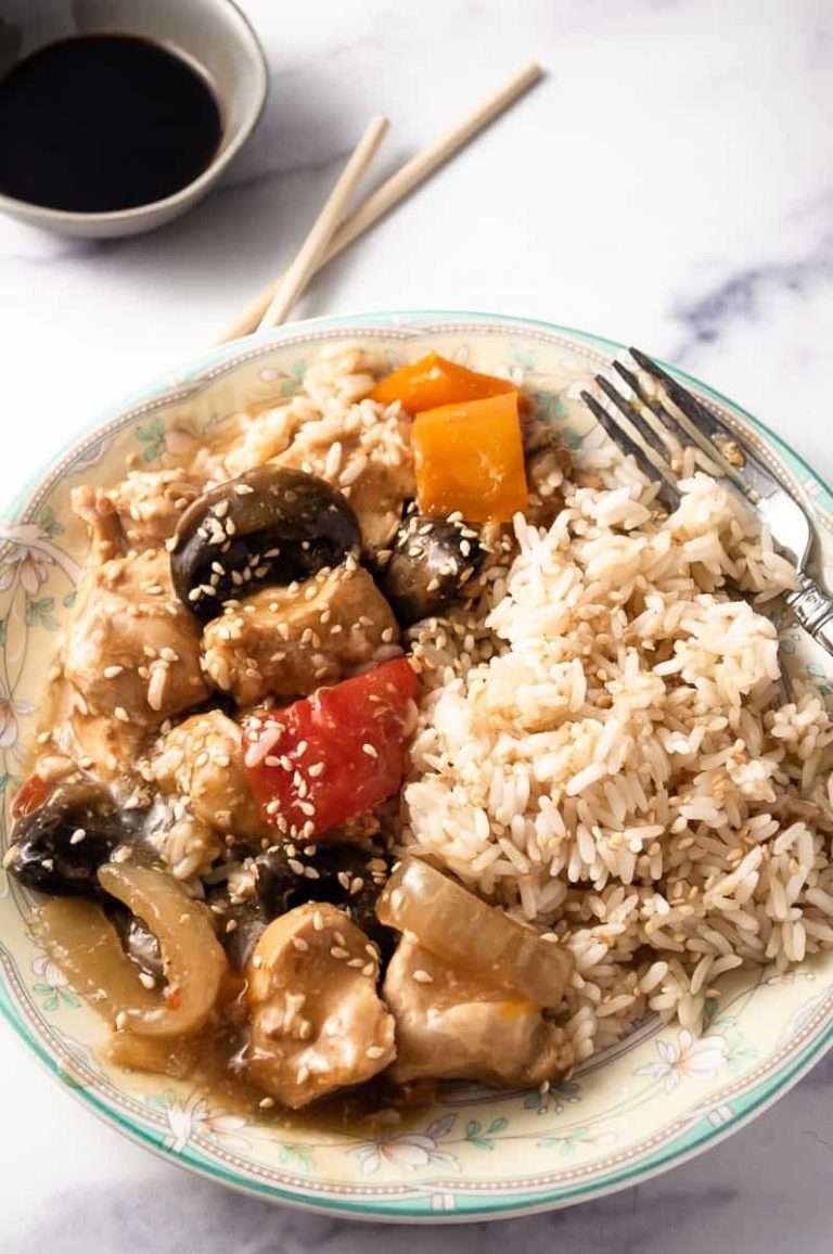 CrockPot Sesame Chicken Easy Recipe on a flowered rim plate with rice a fork chopsticks in the background with a small gray bowl of soy sauce