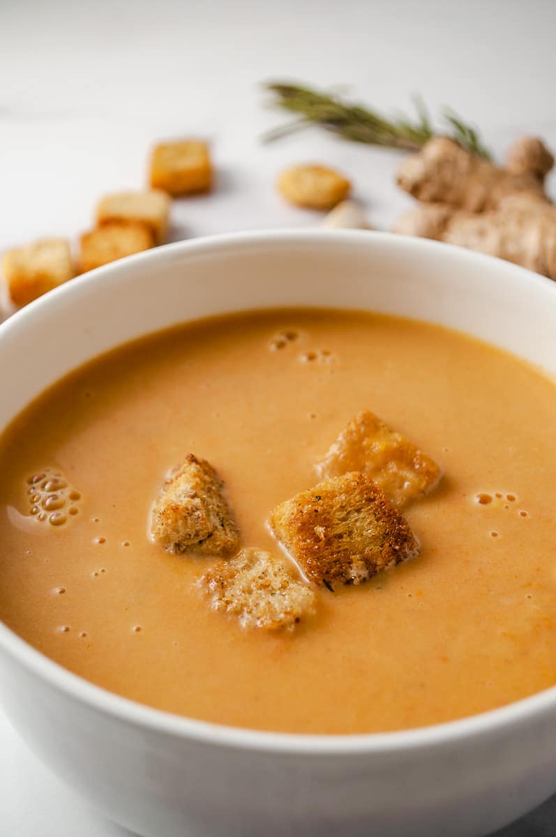 close up of the soup of creamy carrot soup in a white bowl with 4 croutons in the soup, croutons and garlic, ginger root and time on the white counter