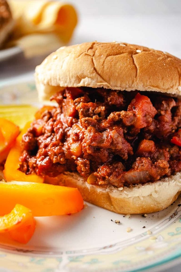 Sloppy jane sandwich in bun with yellow peppers a pickle on the left side and a plate with buns on the top right